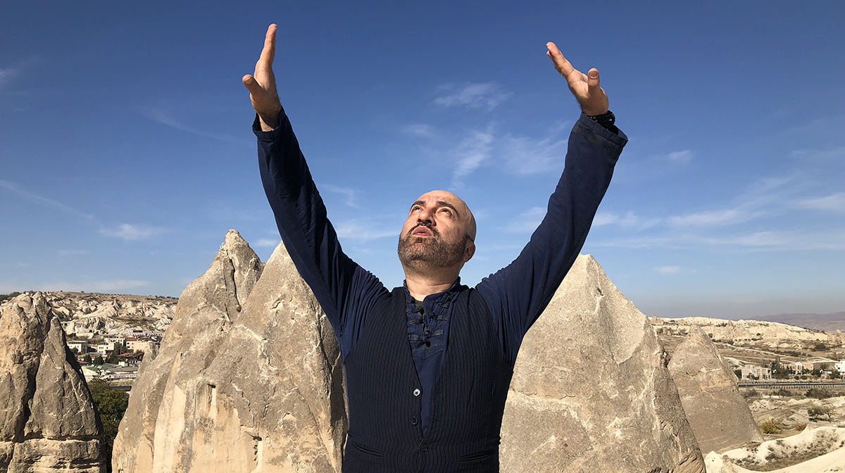 A male performer with arms raised stands outside under a blue sky in front of dramatic rocky peaks.